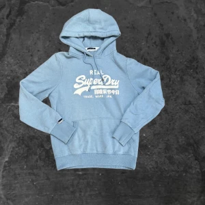 Ljusblå Superdry hoodie - Säljer en ljusblå hoodie från Superdry med stort vitt tryck på bröstet och japanska tecken. Tröjan har huva med snörning, ribbade muddar och en klassisk känguruficka framtill. Perfekt för dig som gillar streetwear och snygga loggor.