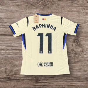 Barcelona Raphinha 11 bortatröja  - Snygg Barcelona bortatröja med Raphinha och nummer 11 på ryggen. Tröjan är ljusgul med svarta och blå detaljer, korta ärmar och tillverkad i lätt funktionsmaterial. Tryckta loggor från Spotify, UNHCR och klubbmärke på bröstet.