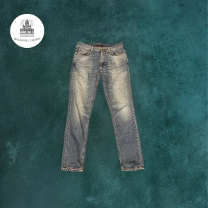 Blå jeans från Nudie Jeans, storlek 33/32 - Snygga blå jeans från Nudie Jeans med klassisk femficksdesign och coola slitningar framtill. Jeansen har raka ben och normal passform, med detaljerade sömmar och Nudie-logga på bakfickan. Perfekt för dig som gillar en avslappnad stil. Har ett litet hål inne i fickan men märks knappt.