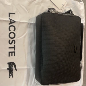 Svart axelväska från Lacoste - Stilren svart axelväska från Lacoste med strukturerad yta och diskret krokodillogo på framsidan. Väskan har rektangulär form, justerbar axelrem och dragkedja upptill. Perfekt för att bära dina essentials med snygg vibe.( Helt ny aldrig använd etiketten osv e kvar )