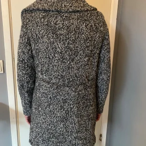Gråmelerad omlottkappa i ullmix - Säljer en gråmelerad kappa med omlottstängning och bred krage. Jackan har lång ärm och är tillverkad i en mjuk ullmix som ger en schysst textur. Perfekt för dig som gillar stilrena och tidlösa plagg med lite edge.