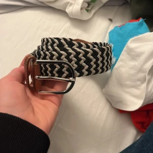 Svart och vit flätat bälte - Snyggt flätat bälte i svart och vitt med bruna skinn-detaljer och silvrigt spänne. Bältet har ett coolt randigt mönster och passar perfekt till jeans eller chinos. Den breda designen ger en modern och trendig look.