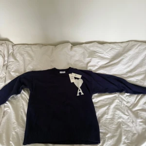 Mörkblå stickad tröja från Ami Paris - Säljer en mörkblå stickad tröja från Ami med ett stort vitt hjärta och bokstaven A på bröstet. Tröjan har rund hals, ribbade muddar och är långärmad. Materialet är mjuk bomull och passformen är som M. Perfekt för dig som gillar stilrena detaljer.