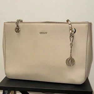 Beige handväska från DKNY med kedja - Super snygg beige/rosa handväska från DKNY med stilren design och guldfärgad kedja som axelrem. Väskan har ett DKNY-emblem i metall, dragkedja upptill och flera fack inuti. Materialet är slätt skinn. Pris går att diskuteras!!!💓