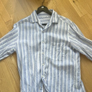 Randig linneskjorta från Loro Piana - Snygg randig skjorta från Loro Piana i 100% linne. Skjortan är vit med blå ränder, har klassisk krage, bröstficka och knäppning framtill. Perfekt för en chill sommarlook med relaxed vibe.