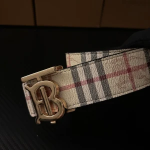Burberry bälte med klassiskt rutmönster - Snyggt Burberry bälte med ikoniskt rutmönster i beige, svart och rött på ena sidan och svart präglat rutmönster med Burberry-ryttare på andra sidan. Bältet har ett stort guldfärgat 'TB'-spänne och är tillverkat i exklusivt skinn.