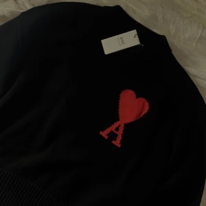 Svart stickad tröja från Ami Paris - Svart stickad tröja från Ami Paris med röd broderad logga i form av ett hjärta och bokstaven A på bröstet. Klassisk rund halsringning och ribbade muddar. Perfekt för dig som vill ha en stilren men ändå unik look.