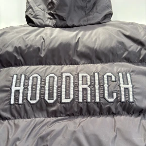 Svart jacka från Hoodrich Junior  - Svart pufferjacka från Hoodrich med stor broderad logga på ryggen och mindre logga på bröstet. Jackan har huva, dragkedja framtill och två sidofickor. Tillverkad i syntetmaterial och perfekt för kalla dagar.