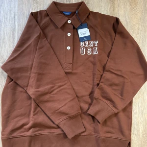 Brun pikétröja från GANT med tryck - Säljer en brun pikétröja från GANT med vit broderad text 'GANT USA' på bröstet. Tröjan har klassisk krage, tre knappar framtill och ribbade muddar vid ärmslut och nederkant. Oversized passform för en avslappnad look.