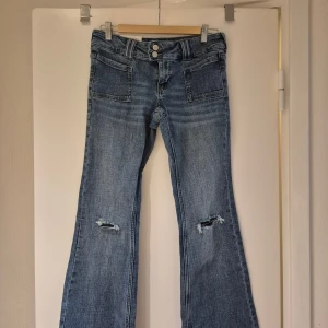 Blå flare jeans low waist från H&M - Säljer ett par blåa flare jeans från H&M i storlek 36. De har låg midja, utsvängda ben och slitna detaljer vid knäna. Jeansen har dubbla knappar i midjan och klassiska fickor fram och bak. Perfekta för en cool och avslappnad stil. Har inte använt de och de har lapparna kvar. 