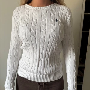 Vit kabelstickad tröja från Ralph Lauren - Vit kabelstickad tröja från Ralph Lauren med klassisk rund halsringning och diskret broderad logga på bröstet. Tröjan har lång ärm, ribbade muddar och en figurnära passform. snygg till allt