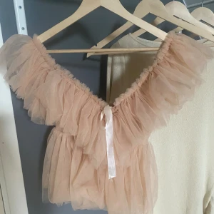 Volang topp - Säljer en fluffig och romantisk beige/rosa topp med volanger och offshoulder-design. Toppen har en söt rosett framtill och är tillverkad i ett tunt, transparent material som ger en drömmig vibe. Perfekt för dig som vill sticka ut med en unik och trendig look. Skriv för fler frågor & funderingar!! 💕💕