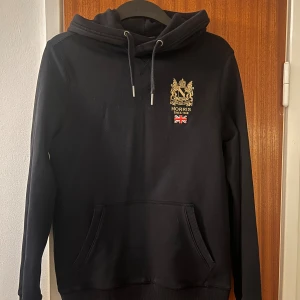 Svart hoodie från Morris med brodyr - Svart hoodie från Morris med guldbroderat emblem och brittisk flagga på bröstet. Klassisk känguruficka, snörning i huvan och mjukt material. Perfekt för dig som gillar stilrena och sportiga plagg med snygga detaljer.