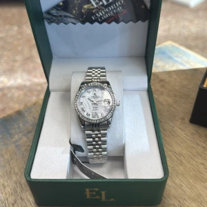 Silverfärgad klocka Emporio Limited - Snygg silverfärgad klocka från Emporio Limited med klassisk länkarmband i metall. Urtavlan är vit med romerska siffror och har en glittrig bezel med stenar runt om. Quartz-urverk och datumvisning. Kommer i grön lyxig box.