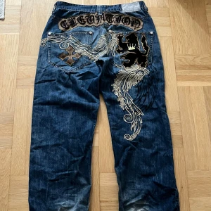 Imperious jeans med broderi och lejon - Säljer ett par blå jeans från Imperious med unika broderade detaljer i guld och svart, inklusive ett stort lejon och texten 'Elevation' på baksidan. Köpta för 1300kr på onlypants61 digitalt kvitto ska nog finnas. Har lite heeldrag längst ner. Hör gärna av dig om du är intresserad!