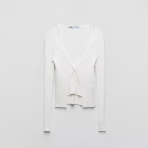 Vit ribbad omlottopp från Zara - Supersnygg vit omlottopp från Zara med ribbad struktur och v-ringning. Långärmad modell med en knapp framtill som ger en stilren och trendig look. Perfekt att styla med jeans eller kjol för en fräsch vibe.