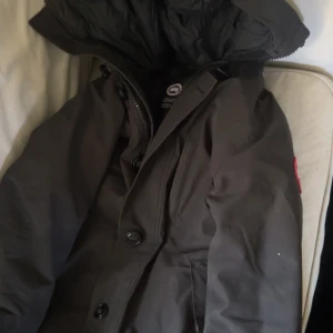 Svart dunjacka från Canada Goose - Säljer en svart dunjacka från Canada Goose med stor huva och klassisk röd logotyp på ärmen. Jackan har både dragkedja och knappar framtill, samt praktiska fickor. Perfekt för kalla dagar och riktigt snygg streetstil. Pris kan diskuteras 