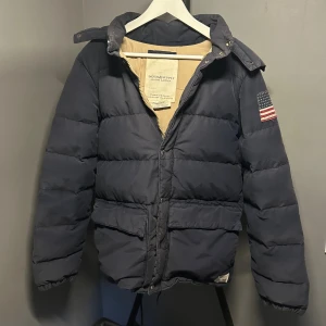 Mörkblå pufferjacka Ralph Lauren - Snygg mörkblå pufferjacka från Denim & Supply Ralph Lauren med patchad amerikansk flagga på ärmen. Jackan har avtagbar huva med fuskpäls, dragkedja och tryckknappar framtill samt stora fickor. Perfekt för kalla dagar. Storlek S men passar mer som medium.