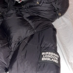 Svart dunjacka från Burberry M - Säljer en svart äkta dunjacka från Burberry med klassiskt rutigt foder i huvan. Jackan har dragkedja, knappar och två fickor med dragkedja. Stort Burberry London England-märke på ärmen. Perfekt för kalla dagar och riktigt snygg streetstil. Och kom med bud gärna.
