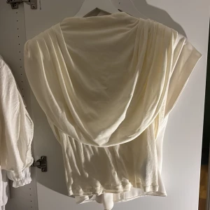 Krämvit bolerotopp från Gina Tricot - Säljer en krämvit bolerotopp från Gina Tricot i mjukt och tunt material. Toppen har en snygg draperad design med korta ärmar och ett elegant fall över axlarna. Perfekt för att addera lager till din outfit och ge en chill vibe.