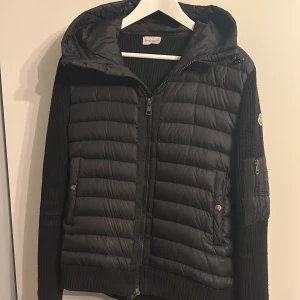 Moncler Cardigan - Svart pufferjacka från Moncler med stickade ärmar och huva. Jackan har dragkedja framtill, två fickor med tryckknappar och en extra ficka med dragkedja på ärmen. Klassisk Moncler-logga på vänster ärm. Perfekt för kyliga dagar.
