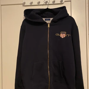 Svart hoodie från GANT med dragkedja - Svart hoodie från GANT med guldfärgad dragkedja och broderad logga på bröstet. Tröjan har huva, fickor framtill och mjukt material som känns skönt mot huden. Perfekt för chill dagar och snygg streetstil.