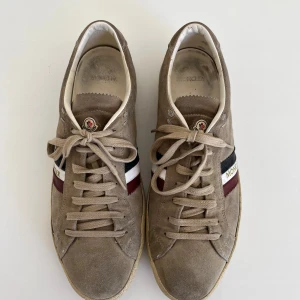 Beige Moncler sneakers i mocka - Snygga beige sneakers från Moncler i mjuk mocka med snörning och rund tå. Sidan har Moncler-logga och ränder i rött, blått och vitt för en cool kontrast. Platt sula och klassisk låg profil gör dem perfekta till jeans eller shorts.