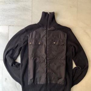 Svart polotröja Julian Adrados - Svart polotröja från Julian Adrados med coola nylonpaneler framtill och två stora fickor med knappar. Stickad i bomull och ull, med hög ribbad krage och dragkedja. Perfekt för dig som gillar street och vill ha något unikt i garderoben.