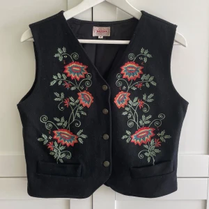 Broderad svart väst  - Svart väst från Indiska med färgglada blombroderier framtill i rött, grönt och blått. 70% ull. Använt men bra skick - inga defekter. 