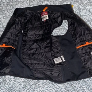Svart dunväst från Parajumpers S - Svart dunväst från Parajumpers i storlek S. Västen har quiltad framsida, dragkedja och en gul detalj vid halsen. Snygg och sportig look med orangea detaljer och lätt glansigt material. Perfekt för lager-på-lager under kyliga dagar.