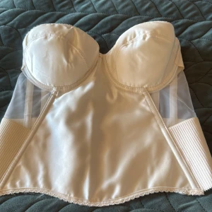 Ivory korsett med mesh och satin - Snygg ivoryfärgad korsett med vadderade kupor, meshpaneler på sidorna och slät satin framtill. Baksidan har breda elastiska band och hakknäppning. Perfekt för att skapa en snygg siluett under klänningar eller toppar.