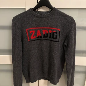 Stickad tröja från Zadig & Voltaire - Snygg grå stickad tröja från Zadig & Voltaire med röd och svart logga framtill. Säljs pga att den tyvärr inte kommer till användning längre