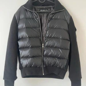 Svart pufferjacka från Bvaldi, stl S - Snygg svart pufferjacka från Bvaldi i storlek S. Jackan har quiltad framsida i glansigt material och stickade ärmar och muddar. Dragkedja framtill och hög krage ger en cool och sportig vibe. Perfekt för kyliga dagar när du vill vara både varm och stilren. Oanvänd.
