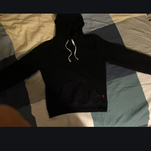 Svart hoodie med vit snörning - Svart hoodie med vit snörning och känguruficka framtill. Klassisk modell med huva och långa ärmar, perfekt för en chill och avslappnad stil. Liten röd detalj vid fickan. Mjuk och skön känsla i materialet.