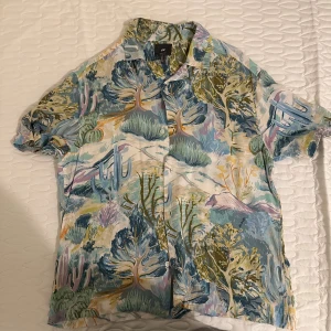 Färgglad kortärmad skjorta från H&M - Kortärmad skjorta från H&M med färgstarkt naturmönster i blått, grönt, lila och beige. Skjortan har klassisk krage, knäppning framtill och loose fit. Perfekt för sommarens varma dagar och ger en chill vibe med sitt unika tryck.