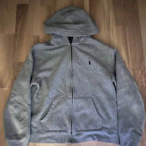 Grå hoodie från Polo Ralph Lauren - Snygg grå hoodie från Polo Ralph Lauren med dragkedja framtill och klassisk huva. Tröjan har två fickor och den ikoniska lilla broderade loggan på bröstet. Perfekt för en chill och stilren look.