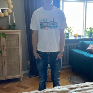 Vit t-shirt med Stockholm-tryck - Vit t-shirt med färgglatt tryck av Stockholmsmotiv på bröstet. Klassisk passform med rund hals och korta ärmar. Perfekt för dig som gillar enkel stil med en touch av stadskänsla. Materialet känns som bomull och t-shirten är lätt att matcha till jeans.