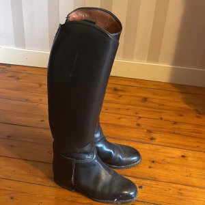 Svarta ridstövlar i skinn - Säljer ett par klassiska svarta ridstövlar med rund tå och platt sula. Stövlarna är tillverkade i slätt skinn med en stilren och hög siluett som går upp till knäet. Insidan är fodrad i ljusbrunt läder för extra komfort. Perfekta för dig som gillar ridning.