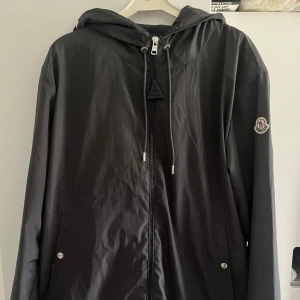 Grå moncler windbreaker  - Mörk grå vindjacka från Moncler med huva, dragkedja framtill och snörning vid halsen. Jackan har två fickor med tryckknappar och Moncler-logga på ärmen. Lätt och stilren design i syntetmaterial, perfekt för blåsiga dagar.