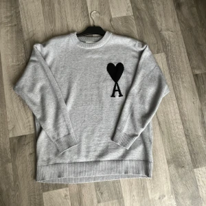 Grå stickad tröja från Ami Paris - Snygg grå stickad tröja från Ami Paris med svart hjärta och A-logga på bröstet. Tröjan har rund hals, ribbade muddar och är långärmad. Perfekt för dig som gillar stilrena och trendiga plagg.