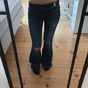 Mörkblå bootcut jeans med slitningar - Säljer ett par mörkblå jeans med bootcut passform och snygga slitningar vid knäna. Jeansen har låg midja, dubbla knappar i linningen och stora fickor både fram och bak. Perfekta för dig som gillar en avslappnad och trendig look.