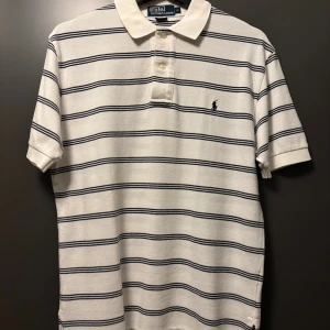 Randig pikétröja från Ralph Lauren - Vit pikétröja från Ralph Lauren med marinblå horisontella ränder och klassisk krage. Tröjan har korta ärmar, knäppning med två knappar och broderad logga på bröstet. Tillverkad i 100% bomull för en skön och luftig känsla.