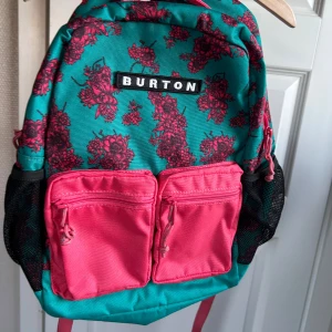 Turkos och rosa rygga från Burton - Snygg ryggsäck från Burton i turkos med rosa blommigt mönster och två rosa frontfickor med dragkedja. Väskan har meshfickor på sidorna, vadderade axelremmar och rymligt huvudfack med innerficka. 