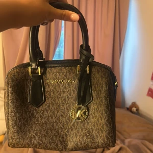 Michael Kors brun handväska med MK-logga - Snygg handväska från Michael Kors i brun och svart med MK-mönster över hela väskan. Väskan har svarta handtag, guldfärgade detaljer och en rund MK-berlock framtill. Materialet är tålig syntet med skinnliknande detaljer och dragkedja upptill. endast använd en gång. 