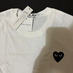 Cdg t shirt - Vit t-shirt från Comme des Garçons Play med det ikoniska svarta hjärtat med ögon broderat på bröstet. Klassisk rund hals och korta ärmar. Tillverkad i 100% bomull för en skön och lätt känsla. Perfekt för dig som gillar stilrena och trendiga plagg.