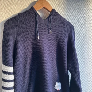 Mörkblå stickad hoodie Thom Browne - Säljer en mörkblå stickad hoodie från Thom Browne med vitrandiga detaljer på ena ärmen. Tröjan har huva med snörning och en tydlig etikett nertill. Perfekt för dig som gillar exklusiva och snygga streetwear-plagg. Jag har inte fått hoodien en så kan inte skicka fler bilder lappen i nacken är borta