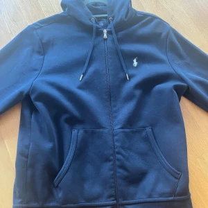 Marinblå hoodie från Polo Ralph Lauren - Snygg marinblå hoodie från Polo Ralph Lauren med dragkedja framtill, huva med snörning och klassisk logga broderad på bröstet. Tröjan har känguruficka och är tillverkad i mjuk bomull, perfekt för chill dagar.