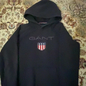 Mörkblå hoodie från GANT med logga - Snygg mörkblå hoodie från GANT med broderad logga och sköld på bröstet. Klassisk huva och ribbade muddar. Perfekt för dig som gillar stilren och sportig look. Passar dig som vill ha en hoodie med premiumkänsla och coola detaljer.