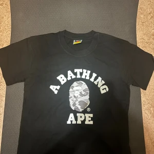 Svart A Bathing Ape t-shirt S - Svart t-shirt från A Bathing Ape med klassisk logotyp och stor camo-apa printad på bröstet. T-shirten har rund hals, korta ärmar och en liten etikett med apans ansikte på ärmen. Materialet är mjuk bomull, perfekt för streetwear.