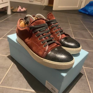 Lanvin sneakers i vinröd mocka - Snygga Lanvin sneakers i vinröd mocka med svart tå i skinn och svarta snören. Klassisk låg modell med vit sula och rund tå. Kommer med originalpåse och box. Perfekta för dig som vill ha en lyxig och stilren sneaker med unika detaljer. Limited edition.